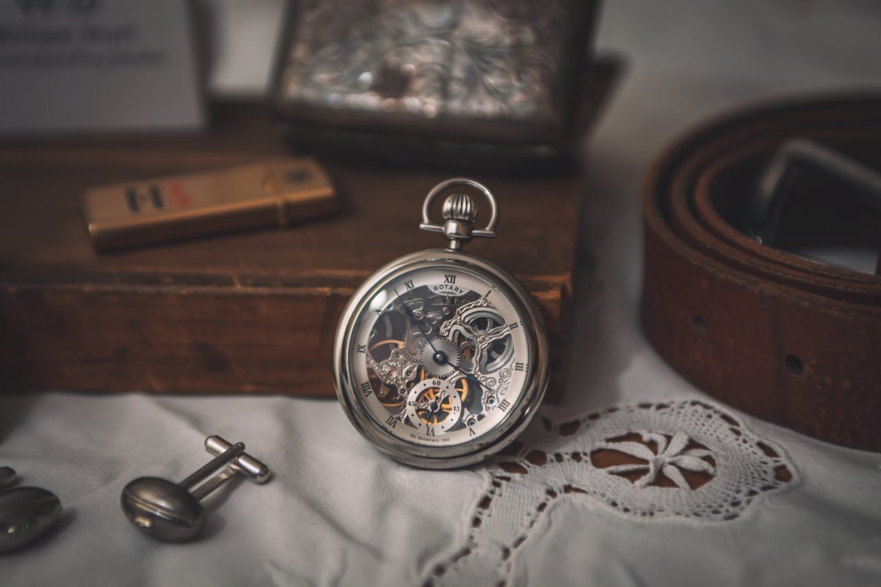 Close-up of a classic skeleton pocket watch on lace fabric, surrounded by a belt and cufflinks.